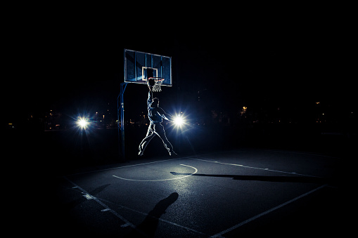 Young man playing basketball at night, going for a slam dunk. Unrecognizable person, male.