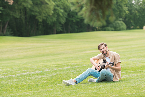 happy young man sitting in green meadow and playing acoustic guitar