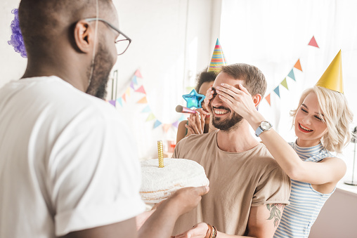 Happy friends covering eyes of young man and greeting him with birthday cake