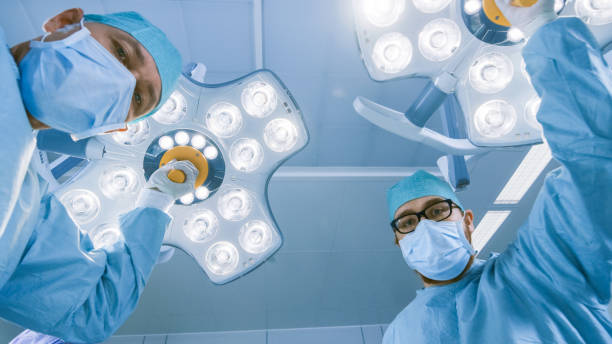 istock Low Angle Shot POV Patient View: Two Professional Surgeons Turning on Surgery Lights while Bending over Patient. 1015934908