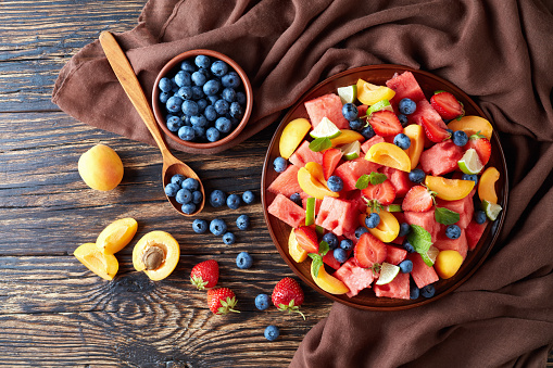 delicious colorful fresh fruit salad with watermelon, blueberries, peach slices, strawberry and lime on a clay plate on an old rustic table with ingredients and napkin, horizontal view from above