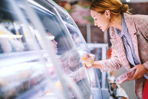 Woman buying food at grocery store. Attractive smiling brunette taking meat from a fridge at local supermarket. Side view. Horizontal. refrigerated section supermarket photos stock pictures, royalty-free photos & images