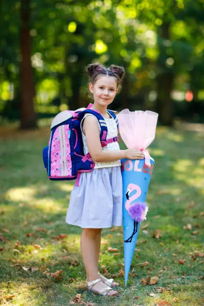 Happy little kid girl with backpack or satchel and big school bag or cone traditional in Germany for the first day of school. Healthy adorable child outdoors, in green park. Elementary class