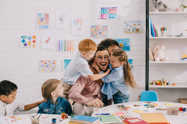 niños interraciales abrazos a maestro feliz en mesa de salón de clases - preschool teacher fotografías e imágenes de stock