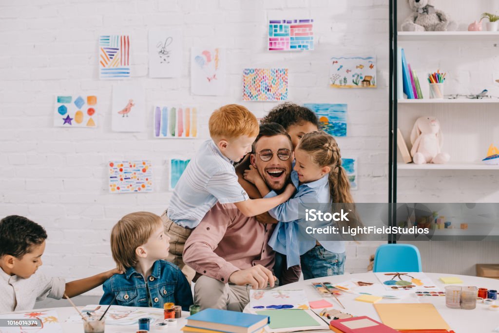 bambini interrazziali abbracciando insegnante felice a tavola in classe - Foto stock royalty-free di Insegnante