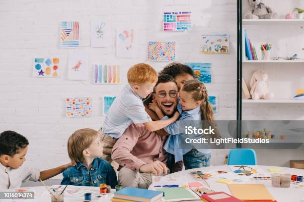 Bambini Interrazziali Abbracciando Insegnante Felice A Tavola In Classe - Fotografie stock e altre immagini di Insegnante