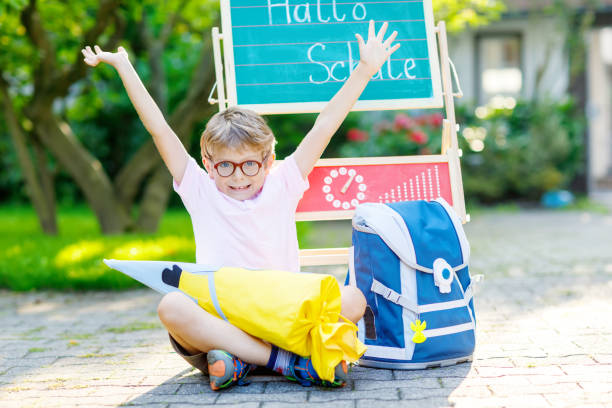 Happy little kid boy with glasses sitting by desk and backpack or satchel Happy little kid boy with glasses sitting by desk and backpack or satchel. Schoolkid with traditional German school bag called Schultuete on his first day to school. Hello school in German language first day of school stock pictures, royalty-free photos & images