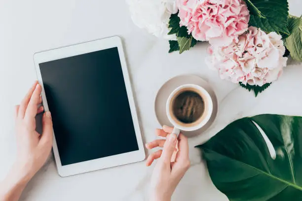 Photo of cropped view of woman hand with coffee cup and digital tablet with blank screen on tabletop with hortensia flowers
