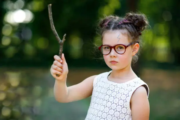 Funny adorable little kid girl with glasses and wooden magic wand playing Harry Potter in park on sunny day. Fan of magic story.