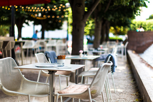 Color image depicting chairs and tables arranged at an outdoors cafe in central London, UK. It is a beautiful green space lined with trees and illuminated with fairy lights. There are bowls of sugar on the tables, but there are no people around. Room for copy space.