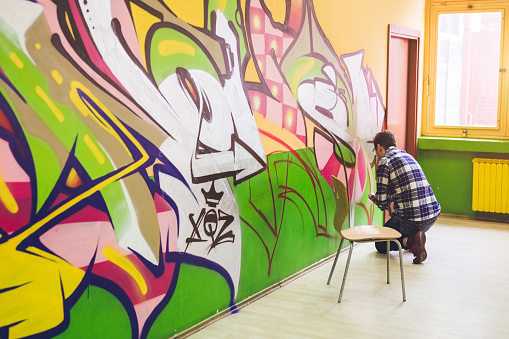 Young man spray painting colorful graffiti on a clean orange wall