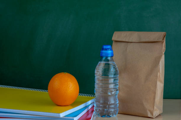 heure du déjeuner. sac à lunch papier brun, orange et bouteille d’eau sur le bureau avec les ordinateurs portables - bag lunch paper bag water bottle photos et images de collection