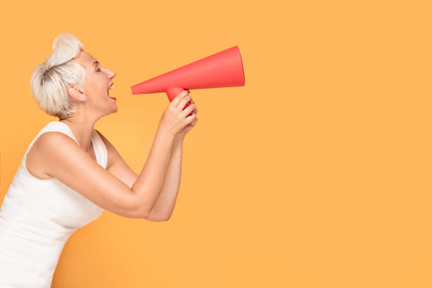 Middle age smiling woman with red megaphone. Middle age elegant woman shouting loud by red megaphone, smiling, looking at camera. Lady expressing positive concept on orange studio background. sold out photos stock pictures, royalty-free photos & images