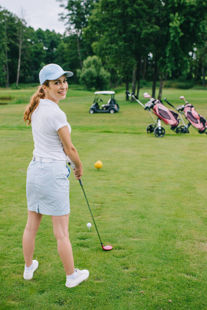 vue latérale du sourire le joueur de golf féminin au cap avec les commandes de club de golf au parcours de golf - golf cart golf mode of transport transportation photos et images de collection