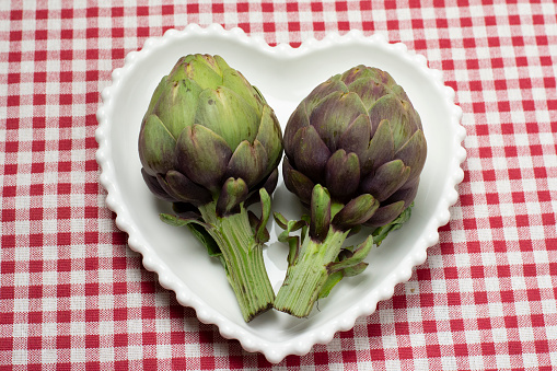 TWO PURPLE FRESH ARTICHOKE ON A HEART SHAPE WHITE PLATE ON  RED AND WHITE CHECKERED TABLECLOTH