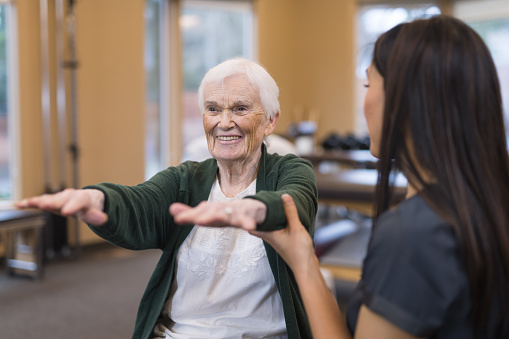 A female occupational therapist of Asian descent works with a cheerful senior female as she does exercises on an exercise ball. She is helping steady her as she holds her arms out.