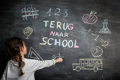 Little girl writing “Terug naar School” multicoloured words blackboard (Back to School in dutch language . She is also drawing a school bus, an apple, a mathematical formula and other school related symbols.