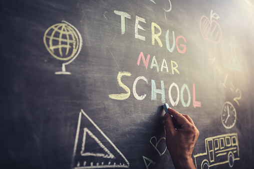 Hand writing “Terug naar School” multicoloured words blackboard (Back to School in dutch language . She is also drawing a school bus, an apple, a mathematical formula and other school related symbols.