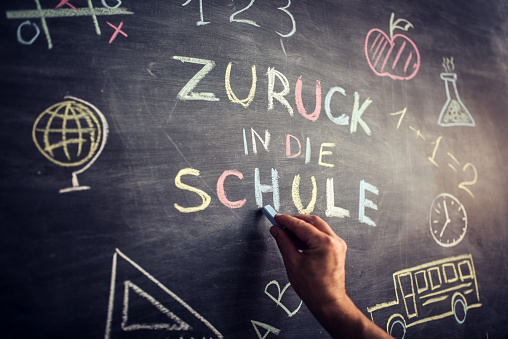 Hand writing “Zuruck in die schule” multicoloured words blackboard (Back to School in german language . She is also drawing a school bus, an apple, a mathematical formula and other school related symbols.