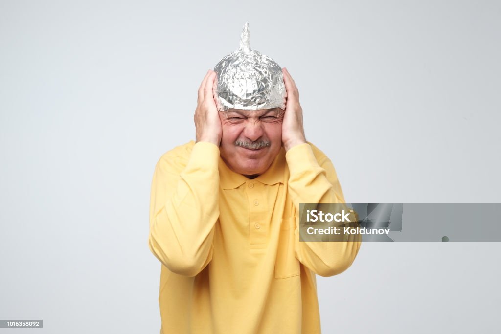 Caucasian mature man in a tin foil hat displeased hiding from outdoor life. Caucasian mature man in a tin foil hat displeased hiding from outdoor life. Afraid of radiation, loud sounds or aliens Aluminum Stock Photo
