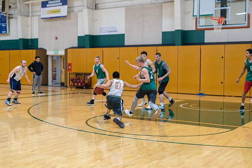 A men's college basketball team practices in the gym. They are scrimmaging and a player in the foreground is driving to the basket while the defense tries to stop him.