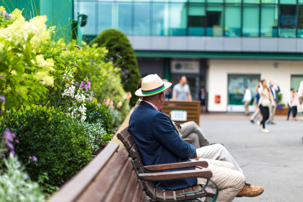 Senior man sitting on bench wearing straw boater hat, Wimbledon, UK Wimbledon, UK - 10 July, 2018: color image depicting a senior man wearing a smart blazer and straw boater hat sitting on a bench in Wimbledon, UK. Focus is on the man, while the background of green foliage and a glass building is defocused in the background. Room for copy space. wimbledon stock pictures, royalty-free photos & images