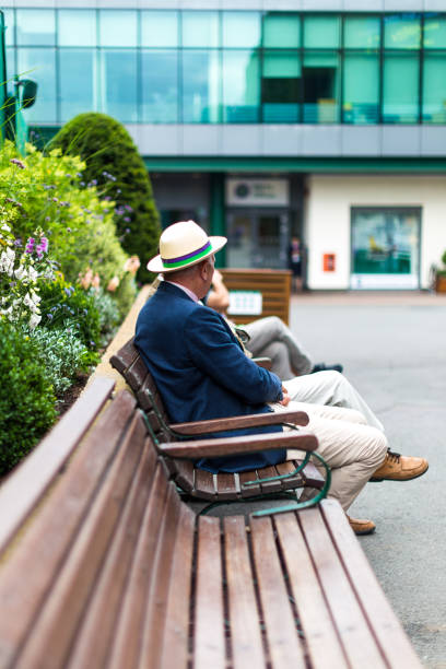 Senior man sitting on bench wearing straw boater hat, Wimbledon, UK Wimbledon, UK - 10 July, 2018: color image depicting a senior man wearing a smart blazer and straw boater hat sitting on a bench in Wimbledon, UK. Focus is on the man, while the background of green foliage and a glass building is defocused in the background. Room for copy space. wimbledon seats stock pictures, royalty-free photos & images
