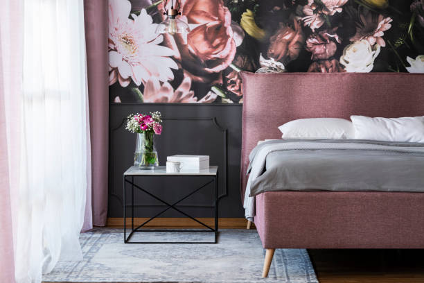 istock Flowers on table next to pink bed with grey sheets in bedroom interior with wallpaper. Real photo 1016513302