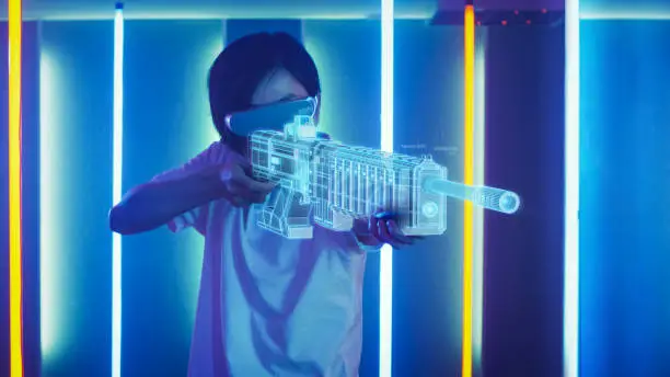 East Asian Pro Gamer Wearing Virtual Reality Headset and Holding Gun Hologram Plays Online Video Game Shooter. Augmented Reality Concept. Cool Neon Colors in the Room.