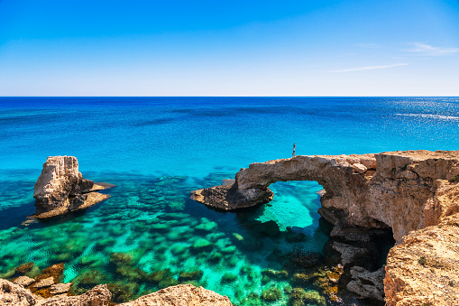 Woman on the beautiful natural rock arch near of Ayia Napa, Cavo Greco and Protaras on Cyprus island, Mediterranean Sea. Legendary bridge lovers. Amazing blue green sea and sunny day.