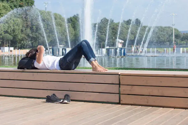 The girl is resting barefoot near the fountain in the park on a hot day. The concept of heat and summer.