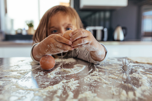 Little girl breaking an egg on kitchen counter covered with flour. Girl child learning cooking in the kitchen at home and making a mess.