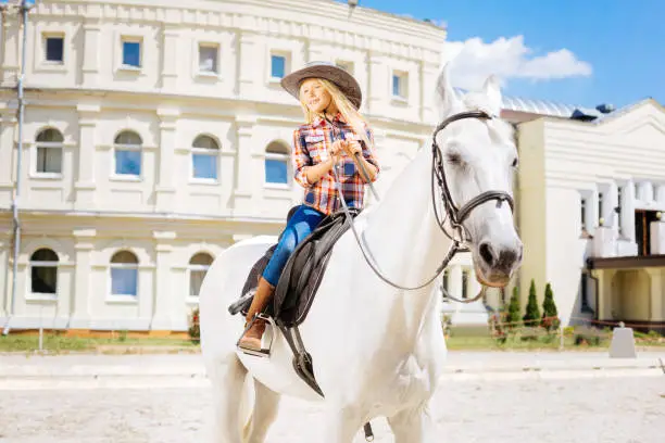 Schoolgirl and equestrianism. Cute schoolgirl wearing stylish cowboy hat fond of equestrianism riding horse on weekend