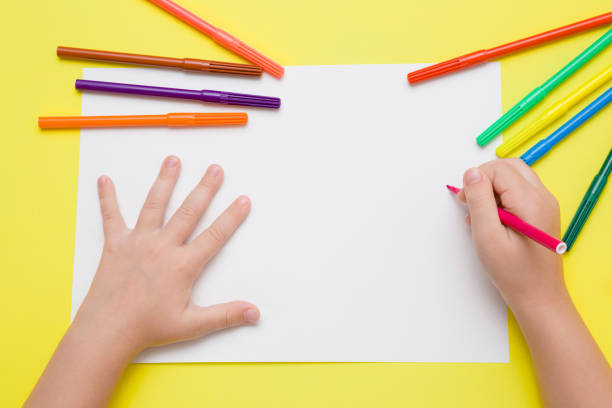 ピンク色のペンと白い紙の上の少女の手の絵。黄色の机の上のカラフルなマーカーです。図面の時に。平面図です。空の場所。 - お絵かき ストックフォトと画像