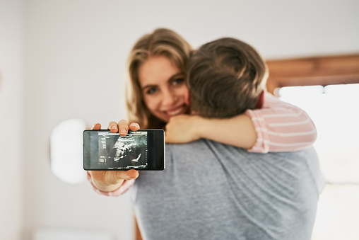Shot of a woman hugging her boyfriend while holding up an image of her sonogram on her cellphone