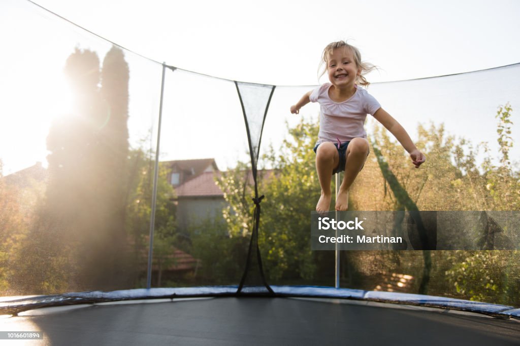 Adorable little girl playing outdoors Cheerful girl in backyard jumping on trampoline Trampoline - Equipment Stock Photo