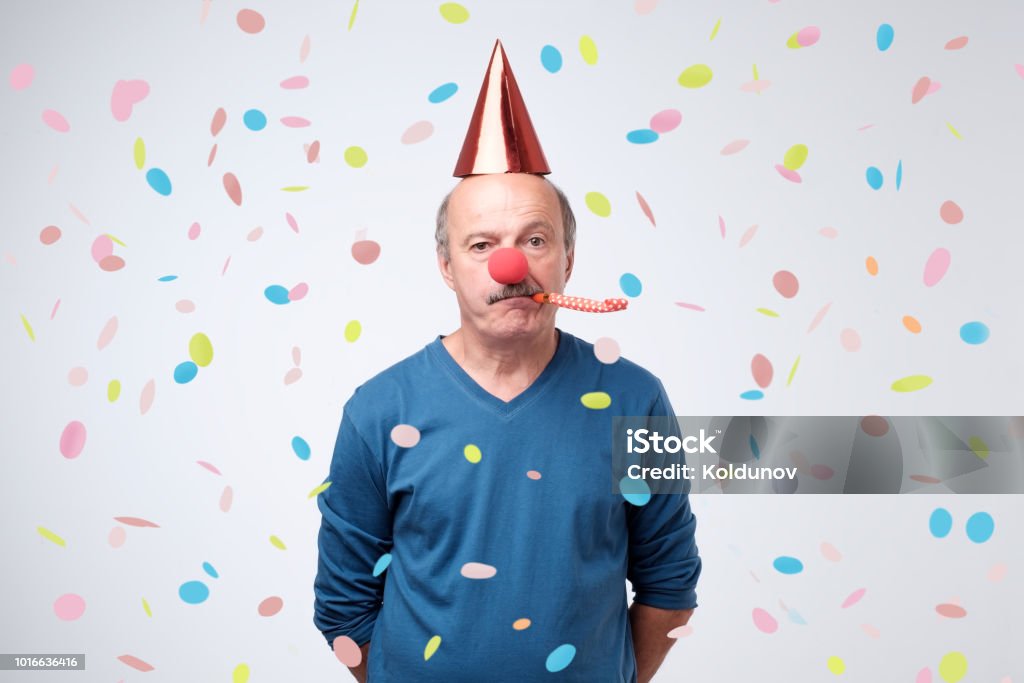 Senior man sad and disappointed because nobody came to celebrate his anniversary. Unhappy birthday senior man is sad and disappointed because nobody came to celebrate his anniversary. Blowing party horn, funny cap and red clown nose on him. confetti flying around him. Sadness Stock Photo