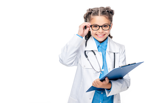 schoolchild in costume of doctor holding clipboard and touching glasses isolated on white