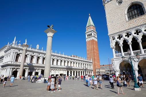 Venice, Italy - August 13, 2017: San Marco bell tower, National Marciana library facade, lion statue and people in the square, clear blue sky in Venice, Italy