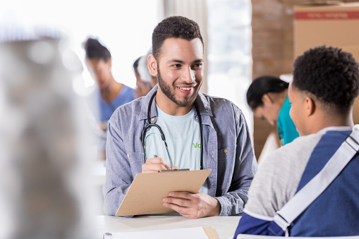 A smiling young male nurse sits at a table across from an unrecognizable male patient.  He holds a clipboard and pen as he listens attentively.