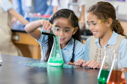Two middle school age little girls smile as they pour a liquid from a beaker to a flask and the chemical reaction causes an overflow.  They are sitting at a table in their school science lab.