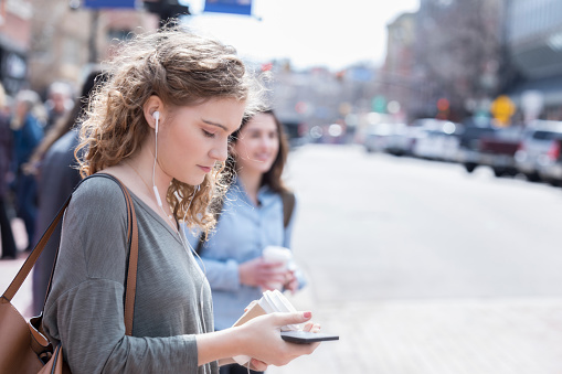 A young woman starts across a city street with two others in the background.  She listens to music on her earbuds as she looks down at her smart phone.