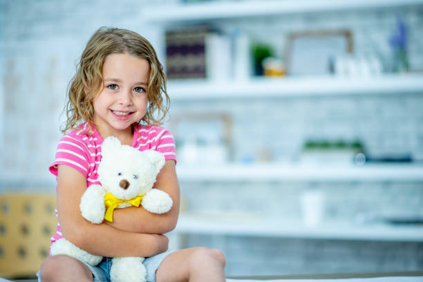 A young girl is indoors in a hospital room. She is smiling while holding a teddy bear to comfort her.