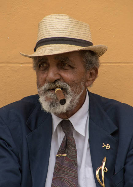 Trinidad, Cuba, Nov 26, 2017 - Old man sits on bench smoking a cigar, against a yellow wall