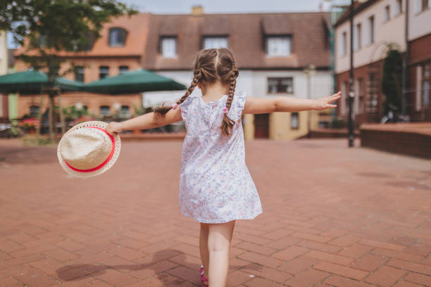 Lovely and beautiful child in a floral dress and a hat, walking on a city square spreading arms.