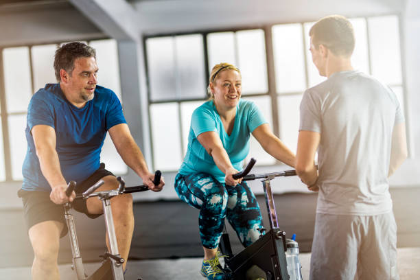 pareja con sobrepeso en las bicis de ejercicio con entrenador personal - gym machine smiling coach fotografías e imágenes de stock
