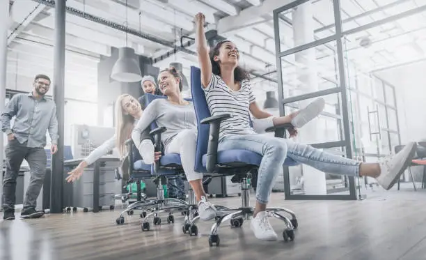 Young cheerful business people dressed in casual clothing are having fun on rowing chairs in a modern office. Happy team concept.