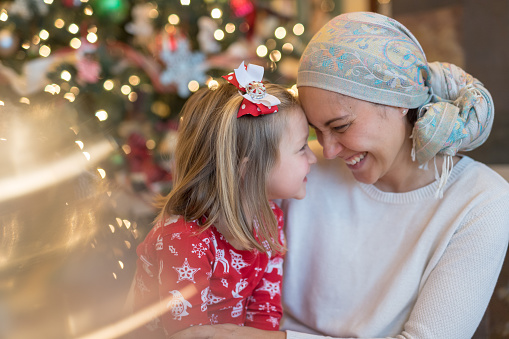 A beautiful ethnic mom wearing a headcovering and battling cancer holds her young daughter playfully on her lap by the Christmas tree in their living room. They're bumping their foreheads together affectionately.