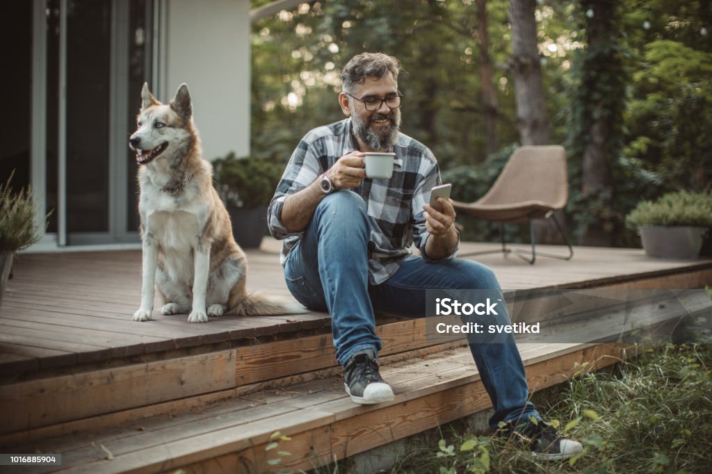 Weekend activities Mature men at his cottage resting on pprch with his dog. Sitting in cahair, drinking coffee and using smart phone. Wearing casual clothing. Men Stock Photo