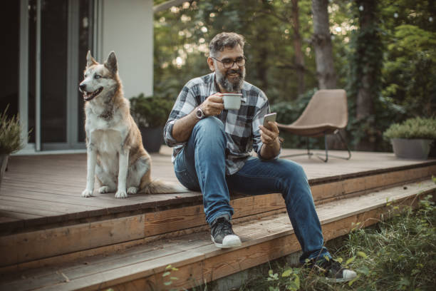 Weekend activities Mature men at his cottage resting on pprch with his dog. Sitting in cahair, drinking coffee and using smart phone. Wearing casual clothing. porch stock pictures, royalty-free photos & images